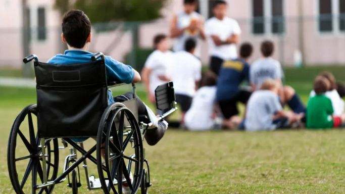 A boy in a wheelchair looks at a group of children sitting on the ground 