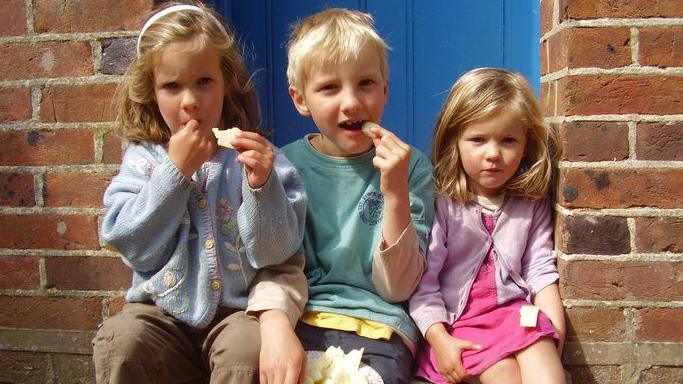 Alex, Will and Mary sit on a step in a doorway together a young children. They are all looking at the camera, eating what looks like some bread. Alex sits on the left and has blonde shoulder length hair and wears a pale blue wool cardigna decorated with embroidered flowers and a pair of beige trousers. Alex is holding a piece of food. Will sits in the middle, he had light blonde short hair and wears a blue short sleeve t shirt with a beige long sleeve t shirt underneath and had a plate of food on his lap. Mary has short blond hair and wears a lilac wool cardigan and pink summer dress. They sit in front of a blue panelled door.