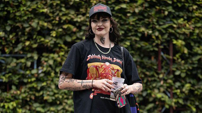A woman stands in front of a hedge. She is wearing a Black Sabbath T-shirt and has tattoos on both arms. She also has a tattoo on her neck and black nail polish. 