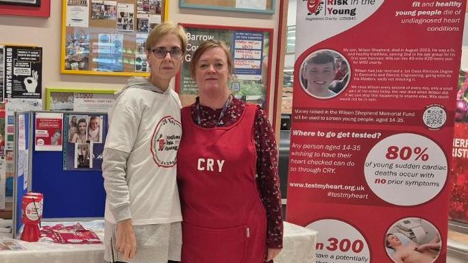 Donna Parker and Lesley Mease stand together in front of charity signage encouraging young people to get their hearts screened. Donna wears a white T-shirt with the charity's logo on, while Lesley wears a red bib with CRY written on it (Cardiac Risk in the Young).