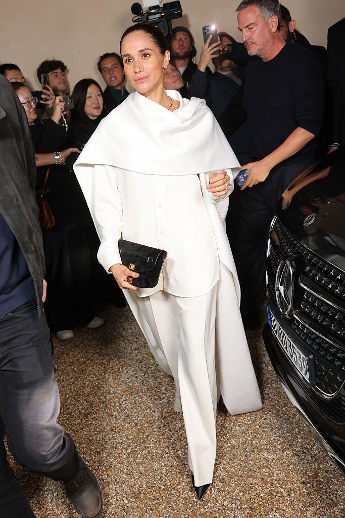 A full length image of the Duchess of Sussex walks in front of a crowd of people dressed in black, several of whom are holding up phones. She is wearing a white jumpsuit with a cape over the shoulders. Her dark hair is tightly tied back.
