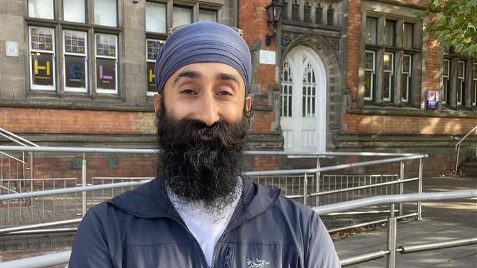 Shuranjeet Singh stands outside the library with his arms folded. He is wearing a blue jacket and blue head covering. He has long, black beard. He is smiling happily at the camera.
