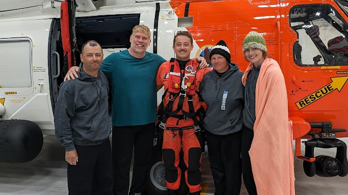 The London's calling rowing team - made up of two men and two women - posing for a photo in front of the US Coastguard helicopter with the pilot who rescued them. One of the women has a blanket around her and the helicopter behind her is red and white.