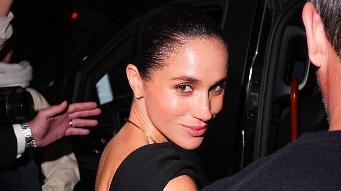 The Duchess of Sussex turns back to look at the camera as she is about to step into a car. A hand can be seen ushering her in. She is wearing a black dress with a gold necklace and stud earrings.