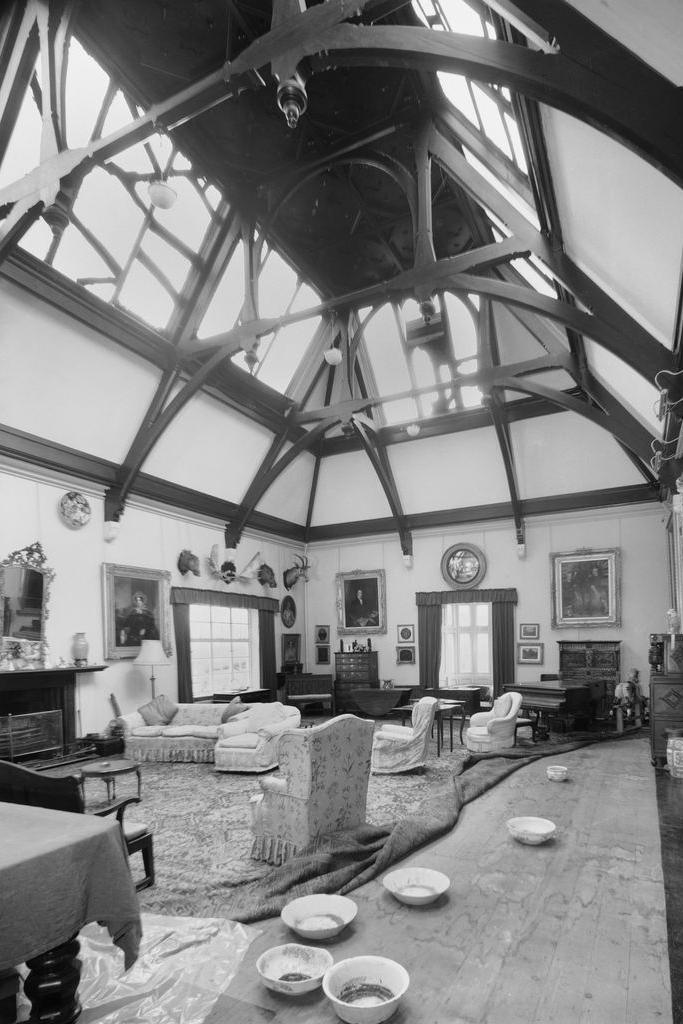 A black and white photo shows furniture including sofas and a wooden table in a large room with a five bay roof. A number of paintings and hunting trophies are hung on the wall. The carpet has been pulled up at one side, with a number of bowls which appear to be there to catch leaks from the roof.