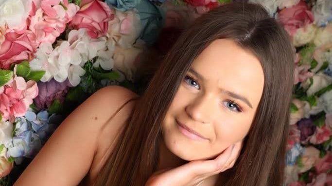 A smiling Amelia Butlin looking directly at the camera. She has blue eyes and dark hair, and is positioned in front of a wall of multi-coloured flowers. 