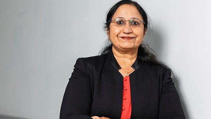 A woman with long dark hair and frameless glasses looks to the camera and is smiling. She is wearing a black suit style top over a red coloured top, and standing in front of a light grey background