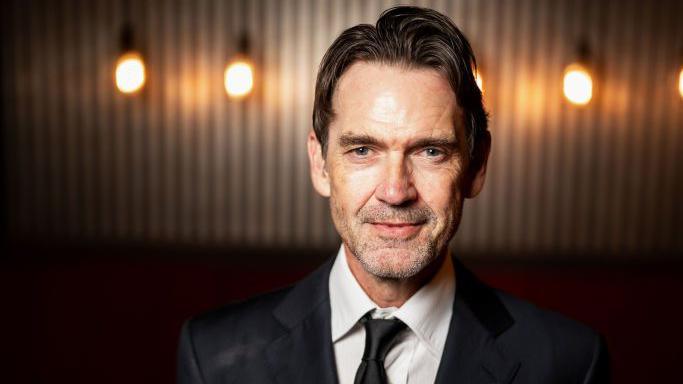 Actor Dougray Scott smiles wearing a black suit and tie and a white shirt. He stands against a dark slatted wall with wall lights.