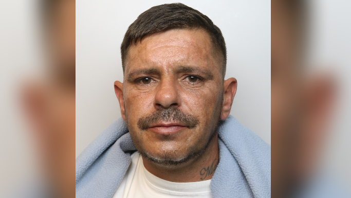 A mugshot of Wolton with short dark hair, a moustache, a baby blue scarf and a tattoo on his neck