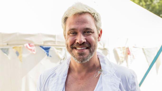 Will Young, a man with light blonde hair and grey stubble and wearing a white shirt, smiling at the camera. There's a white marquee and some bunting behind him and it's a sunny day.