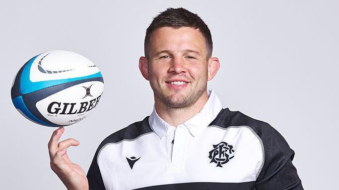 Elliot Dee poses for a camera shoot in a black and white barbarians jersey balancing a rugby ball on the fingers of his right hand