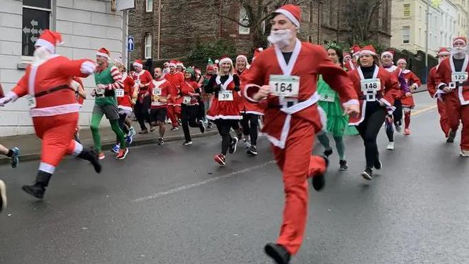 A group of people in Santa and Christmas elf costumes running down a town street in Douglas wearing race registration numbers on their chests. 