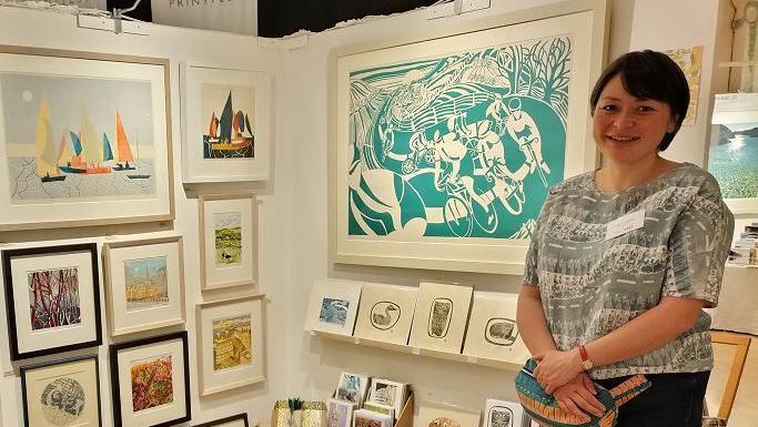 Artists Lisa standing by a display of many paintings and drawings. There are white boards with the artworks all framed. She is standing on the right next to her art, Breeze, it is turquoise and white with cyclists on a windy path surrounded by trees. Lisa herself has short black hair and is wearing a grey Blouse with jeans.