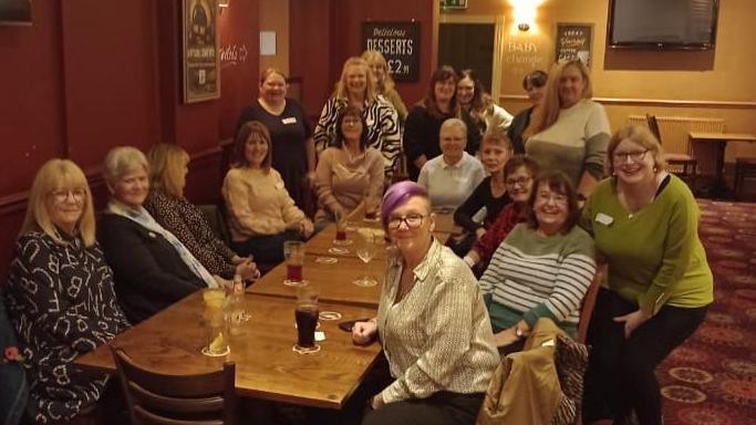 A group of 20 women sit around a long table in a pub. They are looking towards the camera and smiling. 