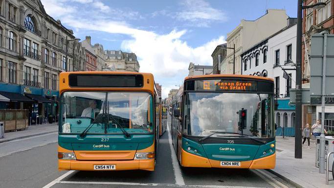 Head on view of a pair of orange and green public service buses in Cardiff city centre. A bus on the right reads "61 Pengam Green via Splott". 