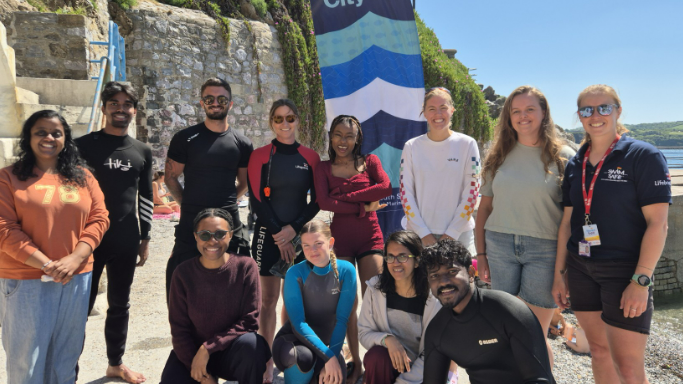 A group of students with instructors on a beach. Four members of the group are squatting on the floor and the rest are standing. Most are in swimwear. Some are in wetsuits. Everyone is smiling.