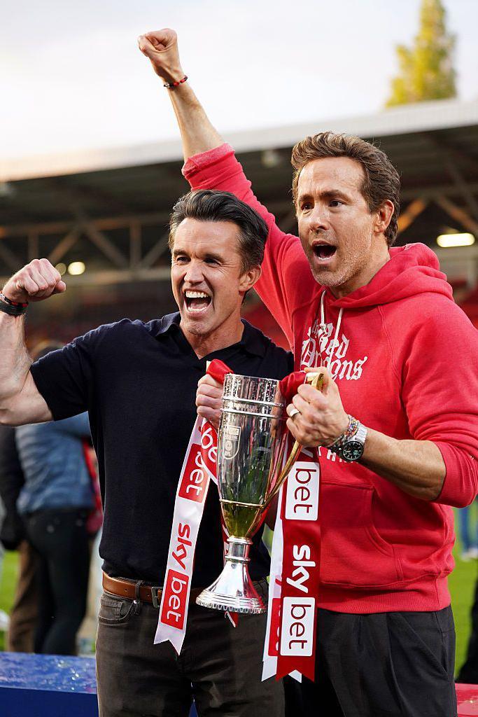 Rob McElhenney and Ryan Reynolds, co-owners of Wrexham AFC, pose for a photo with the trophy following the club's promotion to the Sky Bet Championship. Ryan wears a red hooded jacket, has brown short hair and brown eyes and smiles away from the camera. Rob wears a navy polo shirt and smiles widely at the camera, holding his fist up in the air. They both have one hand holding the trophy. 