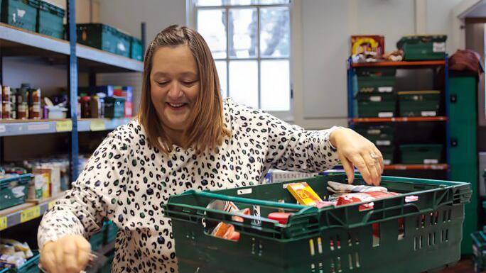 A woman with blonde hair in a black and white spotted top is packing a plastic crate full of food, behind her are shelves of food and a large window