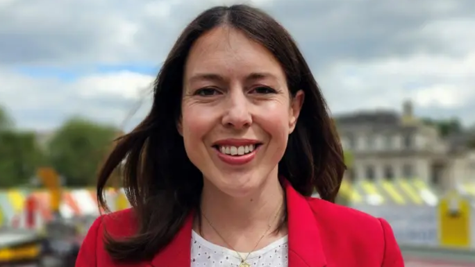 Alice Macdonald is standing outside at the top of Norwich Market, which can be seen in the background behind her. She has brunette hair and is smiling while looking at the camera. She is wearing a white top, gold necklace and red blazer.