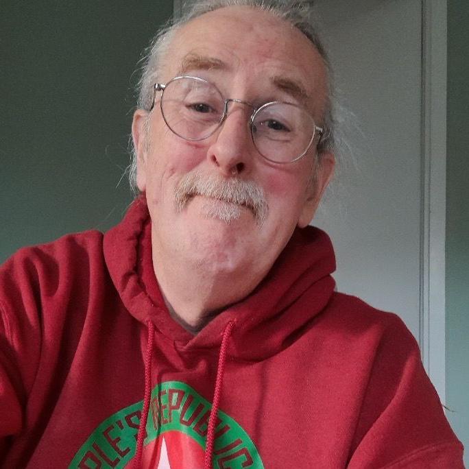 Paul Roberts looking at the camera, half smiling and wearing a red hoodie. He is wearing glasses and his white/grey hair is tied back. He also has a moustache and small goatee.