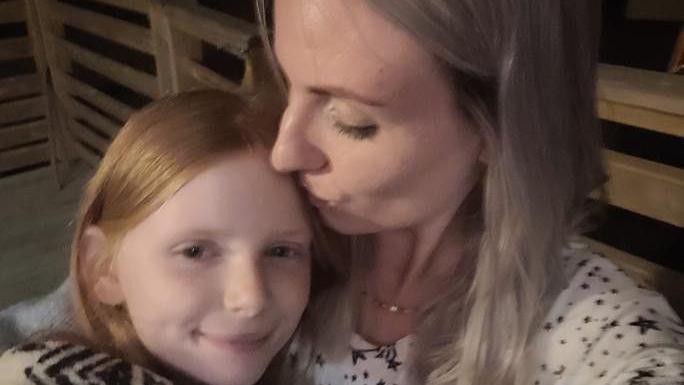 A woman with blonde hair is kissing her young female child on the top of her head. The young girl has ginger hair.