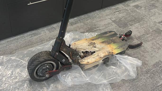 An e-scooter with a burnt foot plate on a corporate, carpeted floor. It sits on top of a sheet of plastic.