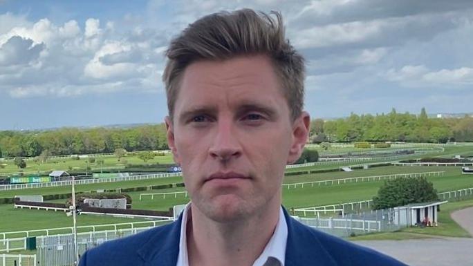A blonde man in a navy suit standing in front of a race course.