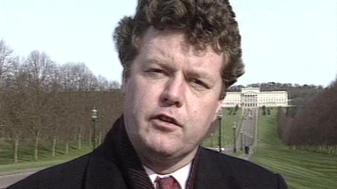 David Davin-Power reporting from Stormont in the 1990s.  He is wearing a black coat, a white shirt, a red tie and a scarf.  He is standing on the long avenue in front of Parliament Buildings.