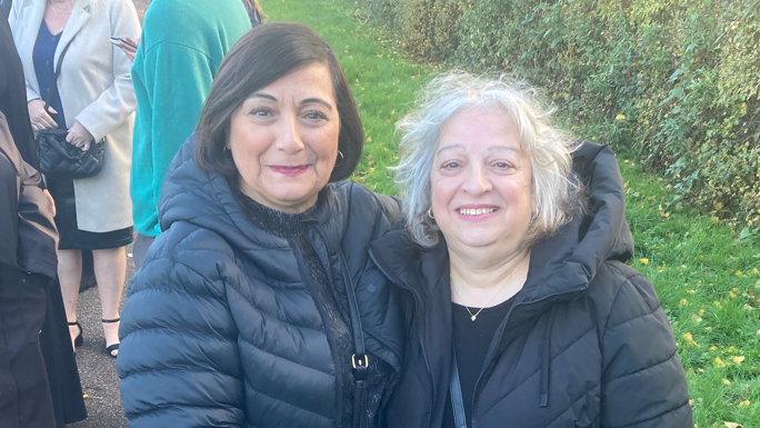 Betty Stefanou was stood on the left was looking towards the camera with a light smile. She had short black hair and was wearing a black coat. On the right was Helen Stefanou who was looking at the camera smiling. She had short grey hair and was wearing a black coat.