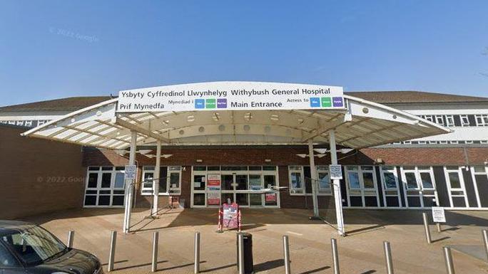 An entrance to a hospital, with a sign at the top and a car parked out front 