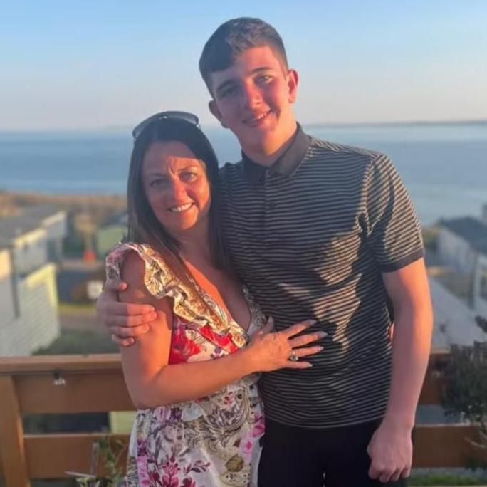 A picture of Wendy and Cole standing on a balcony, with some buildings and the sea in the distance. Wendy is wearing a flowery dress and Cole is wearing a striped grey polo shirt.