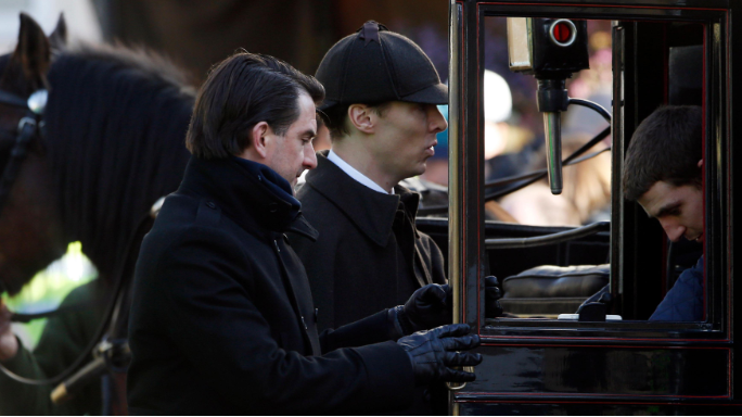 Actor Benedict Cumberbatch is dressed in costume as Sherlock Holmes with a deer stalker cap and dark cloak. He is being shown to a horse-drawn carriage by on-set assistants .
