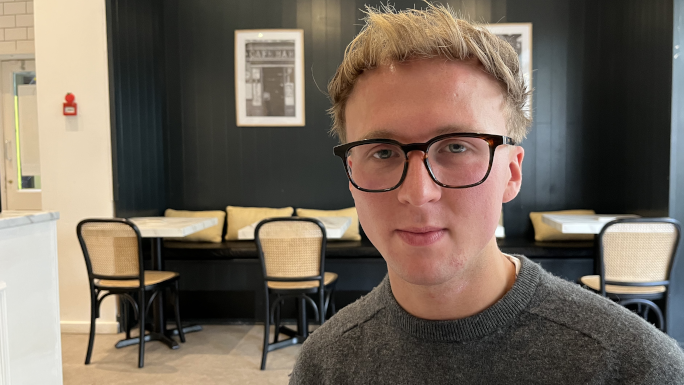 Sam Douglas. He has blonde hair and is wearing a grey jumper and brown rimmed glasses. He is sitting inside Priory Coffee. Behind him are four empty tables.
