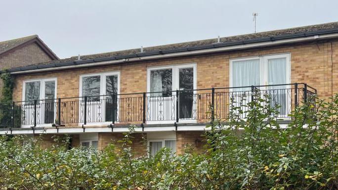 Abbeyfield House is a yellow brick building with a concrete tiled pitched roof. It has white plastic windows and four of the rooms on the upper storeys, which are pictured, feature balconies. There is a large hedge at the front of the property which fronts onto Lawn Avenue.