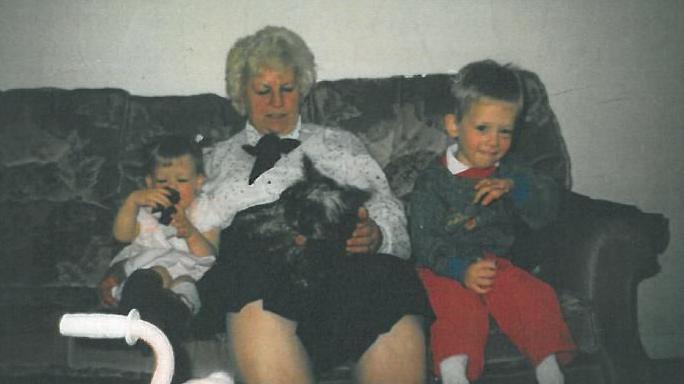 Nora Tait is sat on a sofa in a white top with a small dog on her lap and a young boy on her left and a very young girl on her right