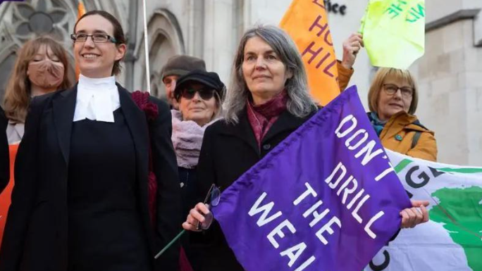 Environmental campaigner Sarah Finch in 2019, having won her court case. She holds a purple flag with Don't Drill the Weald written in white on it, and is standing beside her female lawyer, who is wearing court robes.
Other campaigners stand behind them on the steps of the High Court.