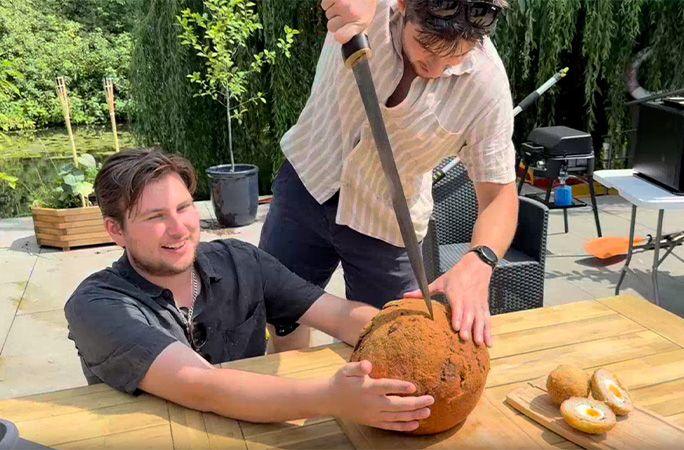 One man with brown hair is kneeling at an outdoor table holding onto a scotch egg that is the same size as a bowling ball. Another man with brown hair holds a sword as he is a bout to cut into the egg.