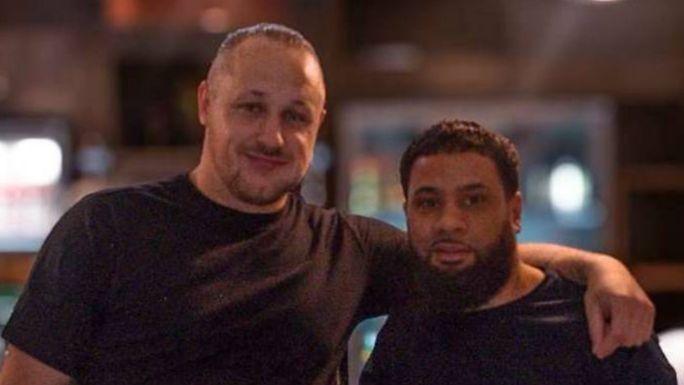 Two men in black t-shirts. On the left a man has his hair tied back, the man on the right has dark hair and a thick, dark beard