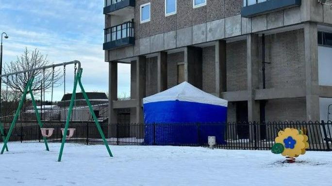 Scene of man's death, snow on grounds, high-rise in background, blue and white police tent, and the green frame of swings for children.