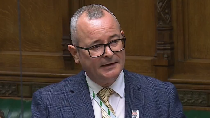 Lee Barron wearing a navy blue blazer, white shirt and yellow and black tie speaking in the House of Commons. 