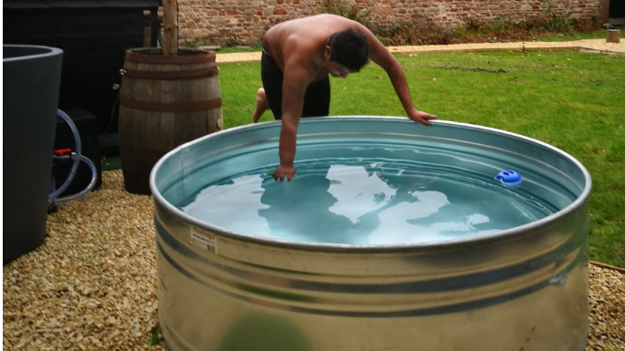 A man wearing shorts carefully tests the temperature of an ice bath. It is a large stainless steel bath, very nearly full of water. It stands on a patch of gravel in front of a grassy lawn. 