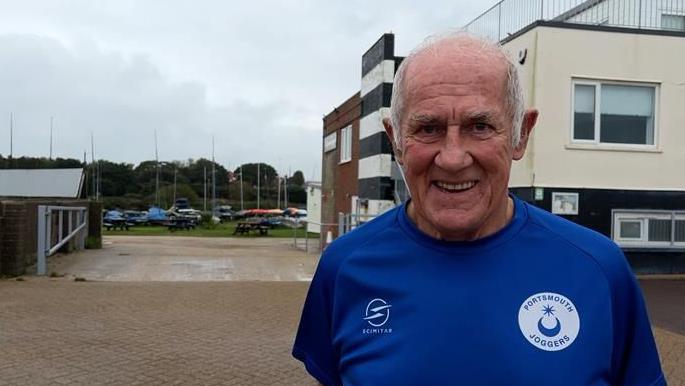 Phil Pollard smiling for the camera on the street on a cloudy day. He is wearing a dark blue shirt branded "Portsmouth Joggers"