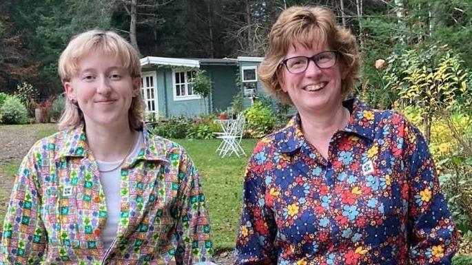 Eilidh and her mum Jenny. Eilidh has blonde shoulder length hair and she is wearing patterned pink, green, yellow and purple dungarees. She is stood in a garden with a summer house in the background. Jenny is smiling beside her wearing blue, yellow and red floral patterned dungarees. She is wearing glasses and has brown hair.