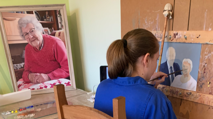 A woman in a blue shirt with her back to the camera, paints two people on canvas. A portrait of her grandmother stands to her left 