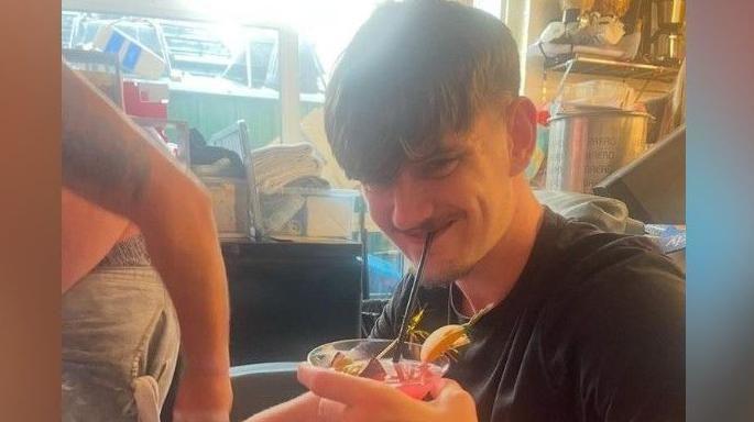 Tyler Kerry has short dark hair. He is smiling at the camera while sipping a pink cocktail through a straw. He is wearing a black T-shirt. 