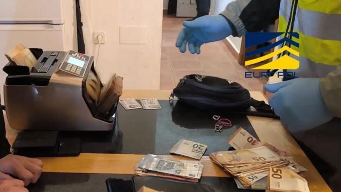 The hands of two investigators examining a large pile of euro notes on a table. There is also a money counter. The image is watermarked with the logos of the Spanish National Police and Interpol. 