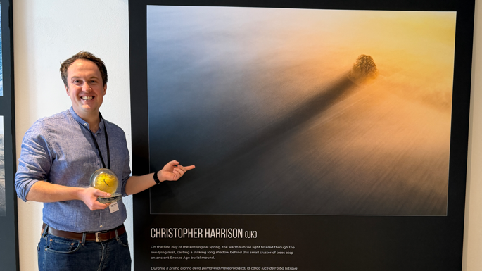 A smiling Christopher Harrison pointing at his photograph in an exhibition. He is holding his award.
