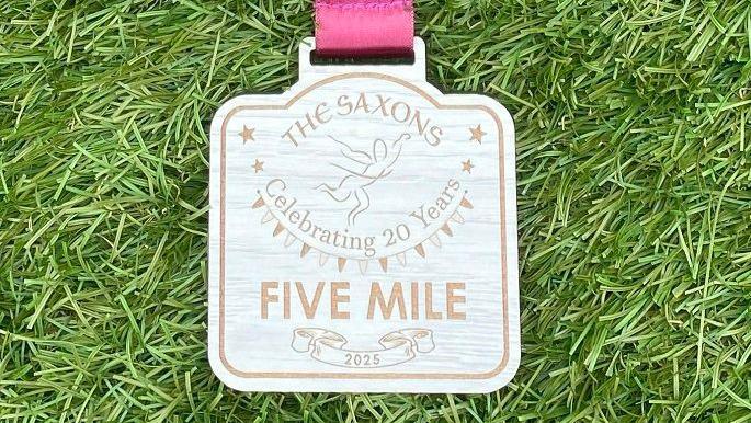 A medal that has been engraved and reads: The Saxons, celebrating 20 years, five mile, 2025. It sits face up on grass. 