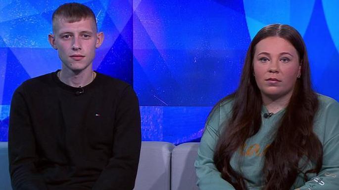 A man and a woman sit side-by-side on a sofa. The man, on the left, is wearing a black top, and the woman on the right has long flowing dark hair which runs over her jumper.