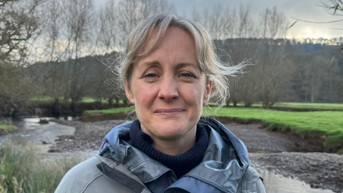 Solicitor Penny Gane standing by the River Onny in Shropshire. She has light blonde hair, pulled back behind her head. Behind her, the waterway runs between green, grassy banks and grey pebbles on the other side. Behind the waterway are several trees. Ms Gane wears a grey jacket with "Fish Legal" on it in white letters and the jacket has a blue/grey collar. The collar of a blue jumper can just be seen underne